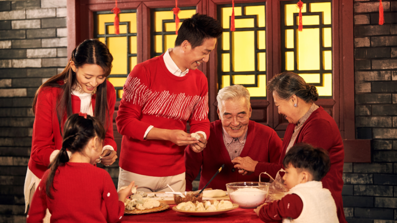 Большая китайская семья в красных свитерах вместе готовит традиционные пельмени за столом.